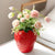 Vase rouge en forme de fraise avec bouquet de fleurs posé sur une table blanche.