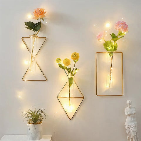 Soliflore cadre mural géométrique en verre et fer forgé présentation des modèles sablier, prismatique, rectangulaire avec fleurs et guirlande lumineuse accrochés sur un mur.