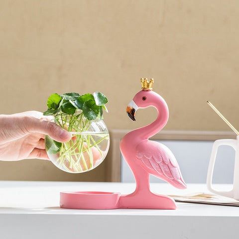 Soliflore Flamant Rose Couronné présentation du modèle petit 1 avec plantes vertes sur une table blanche, contenant en verre tenu en main.