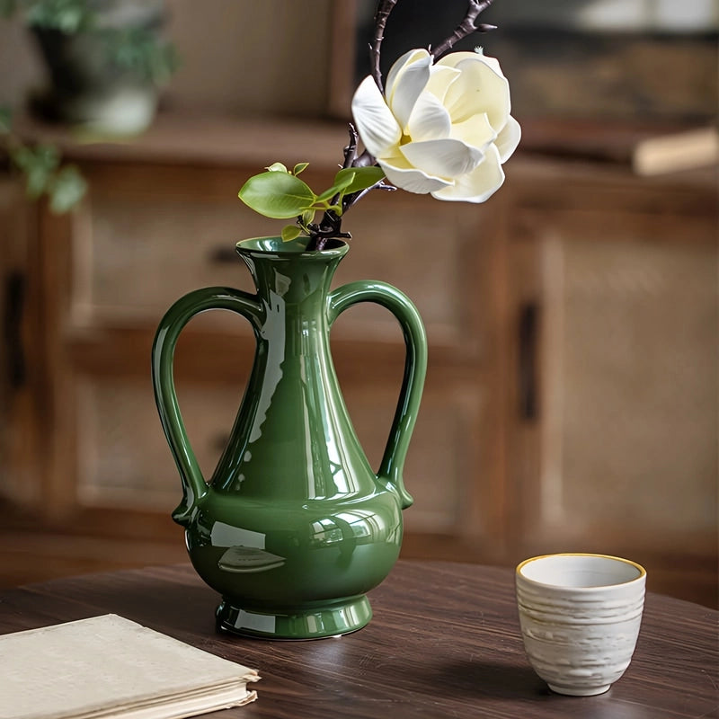 Vase Amphore Verte à Double Anse, présentation avec une tige de fleur blanche sur une table en bois.