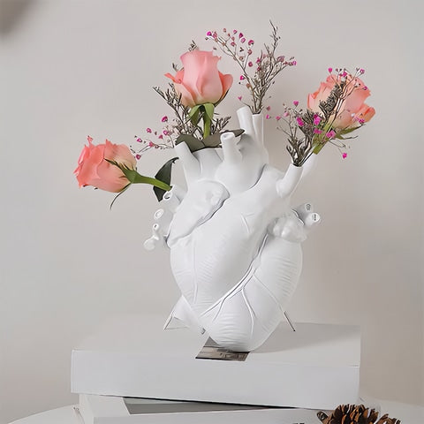 Vase cœur humain blanc en céramique avec roses et fleurs posé sur une pile de livres, sur une table.