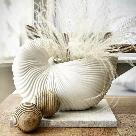 Vase Coquillage Blanc en céramique présentation avec plumes blanches et ornements sur une table en bois.