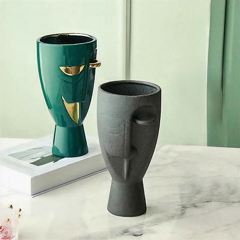 Vase en forme de visage style nordique mise en scène des modèles Vert Doré sur un livre et Noir Moucheté posé sur une table.