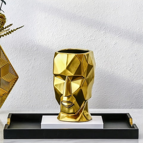 Vase visage design doré en céramique présentation sans fleurs posé sur un livre dans un plateau, le tout sur une table.
