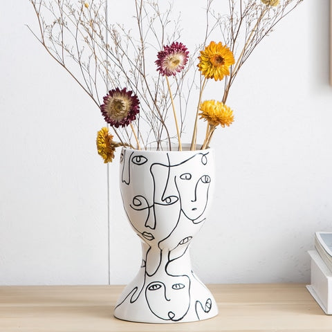 Vase visages abstraits original en céramique présentation avec fleurs séchées sur un meuble en bois.