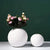 Deux vases boule blanche avec fleurs posés sur une table blanche.