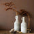 Deux vases en forme de bouteille blanc avec fleurs séchées posés sur une table en bois.