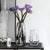 Deux vases en verre avec bouquet de fleurs violet posés sur un meuble noir.