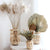Plusieurs vases en bambou avec fleurs séchées posés sur une table blanche.