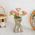 Vase argenté difforme avec bouquet de fleurs posé sur une table blanche.