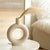 Vase donut beige avec fleurs séchées posé sur une petite table ronde blanche.