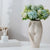 Vase fessier de femme blanc avec bouquet de fleurs posé sur une table blanche